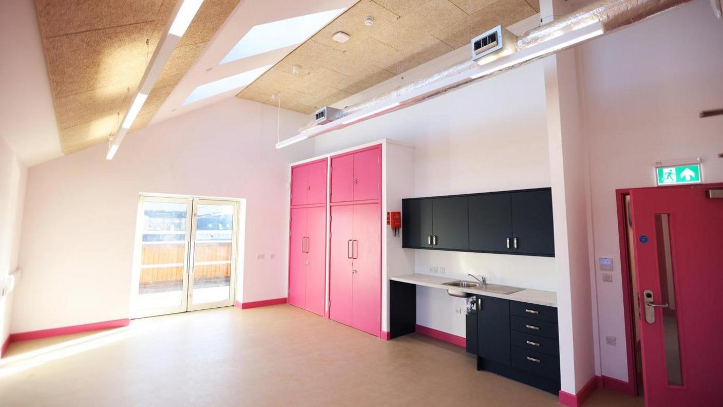 A large and bright room has sunlights in the roof and glass double doors to the outside.  It is painted white, with a light couloured floor and pink cupboards, interior door and skirting boards.  There is a metalic sink with black cupboards and drawers.