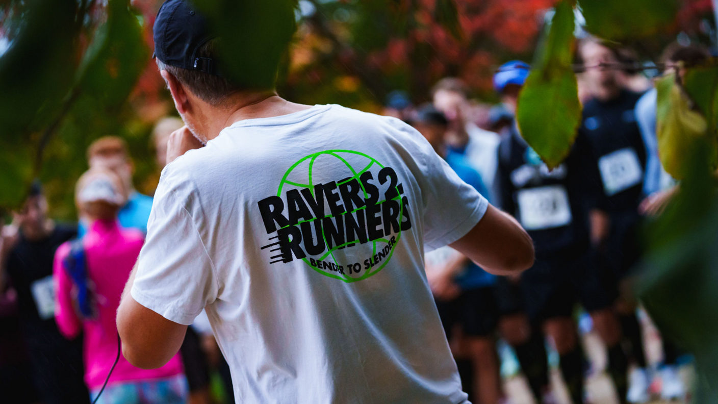 A man in a white t-shirt seen from behind, with the logo Ravers2Runners across the back of his shirt. A group of runners are blurred in the background.