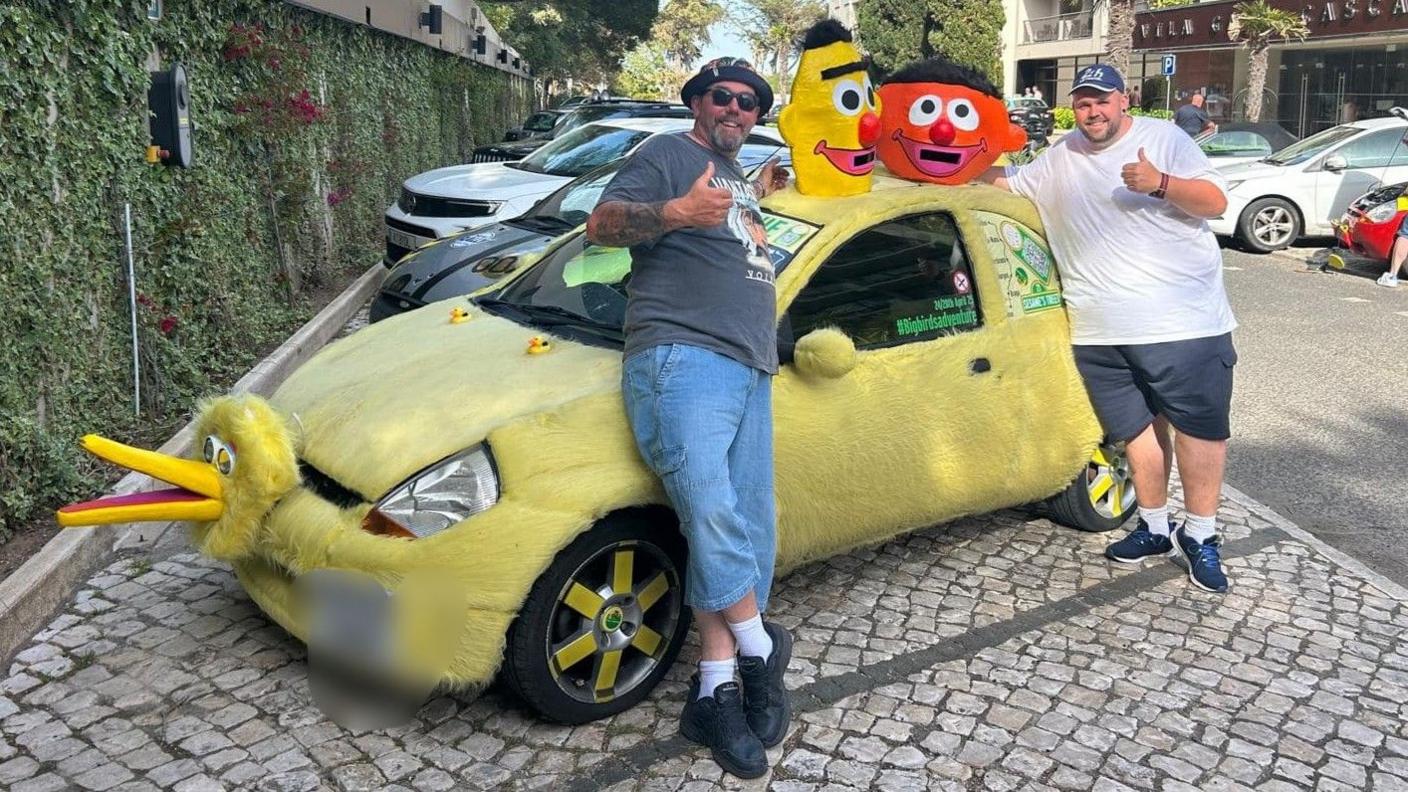 A furry yellow car decorated to look like Big Bird from Sesame Street is parked in a European street. Two men wearing shorts and T-shirts are standing next to it, giving the thumbs-up and smiling.