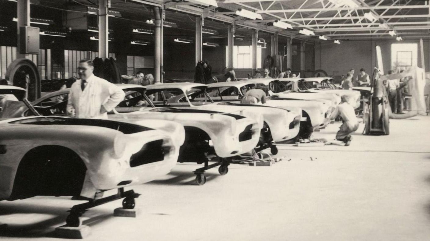 This is a black and white image that shows the shells of a series of cars lined up in a large industrial unit. A man in a white coat is looking at the car closest to the camera. People can be seen working on the cars in the background.