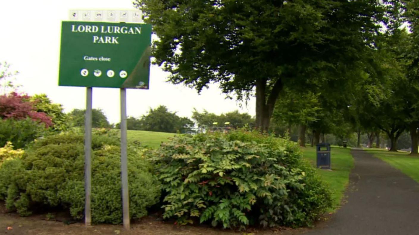 A sign saying Lord Lurgan Park. There are bushes, and trees in view and a pathway.