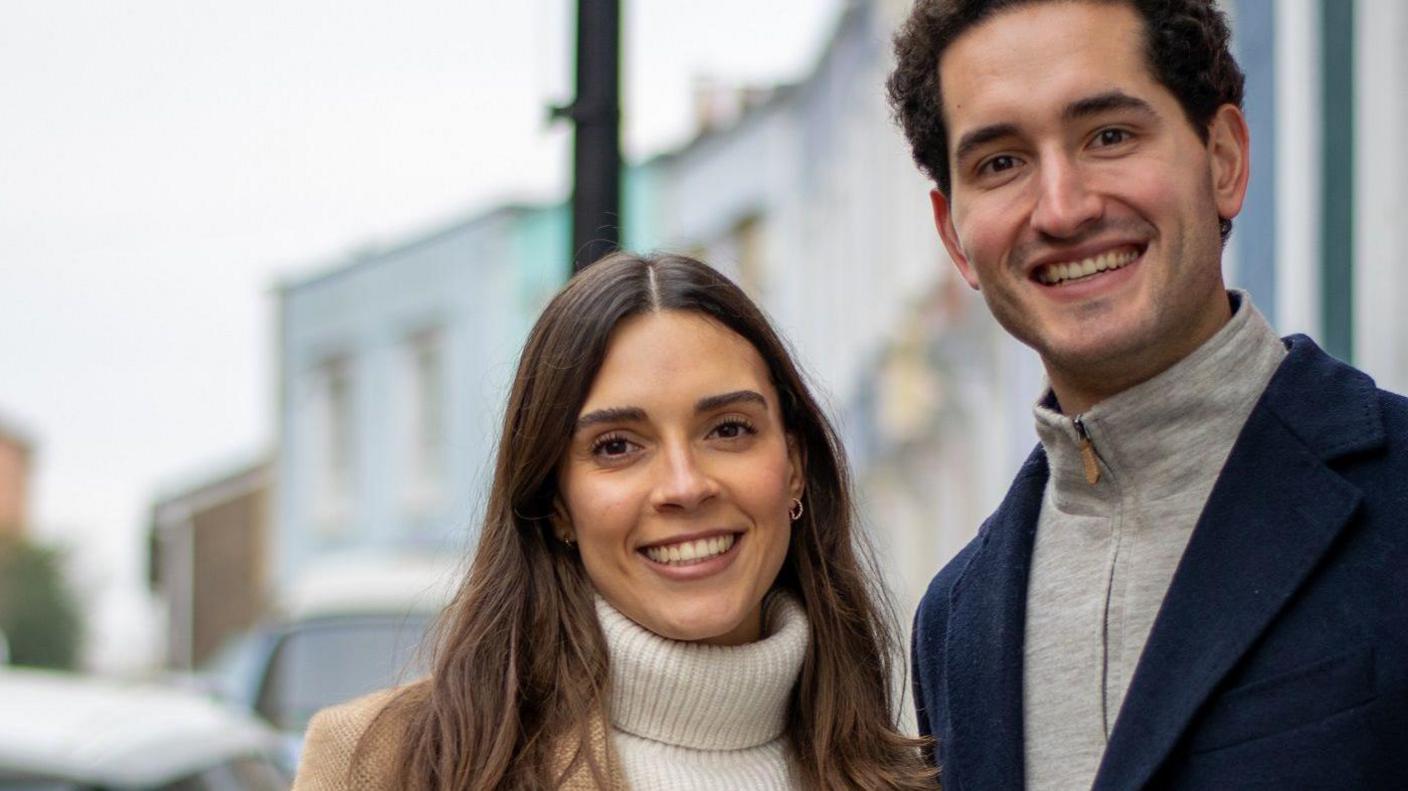 Close up of couple smiling. She is wearing a white jumper and he is wearing a grey top. They are stood in front of houses which are blurred in the background.