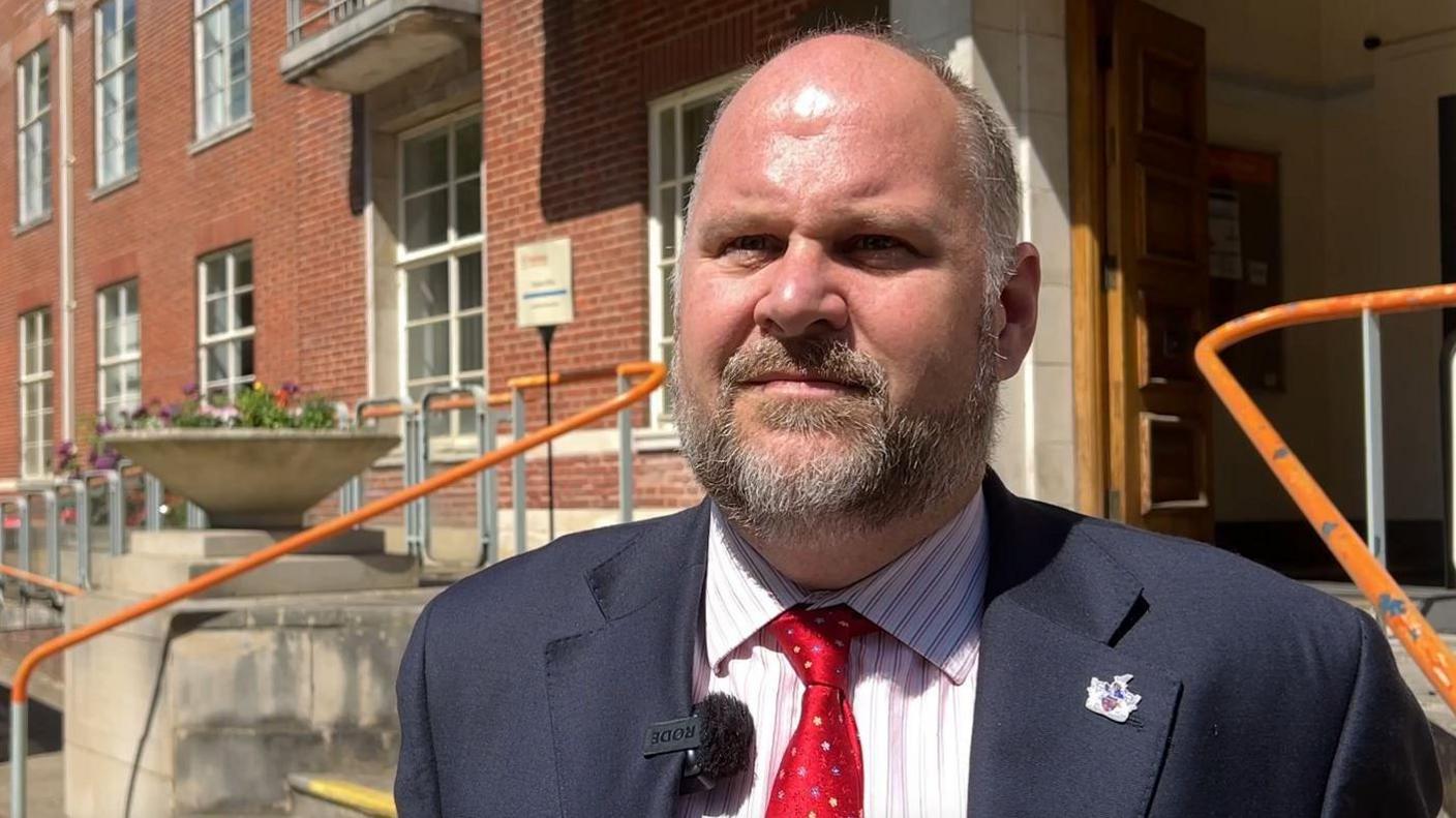Jim Robbins outside red brick council building looking at the camera. He wears a suit with a red tie.