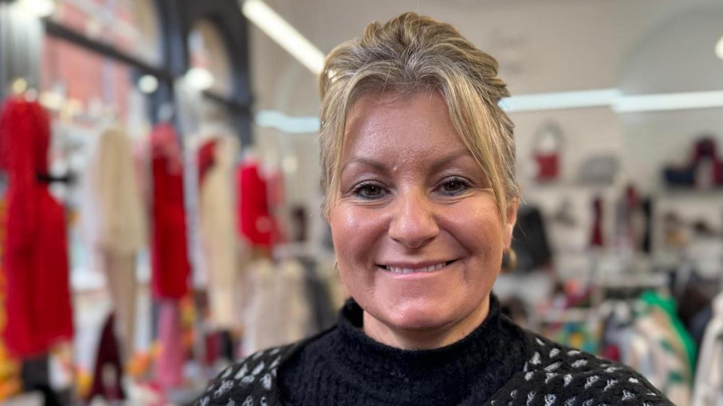 Joanne Holderness smiles as she stands in her clothes shop and looks into the camera. She has blonde hair, tied back, and wears a black polo-neck top and a black and white cardigan.