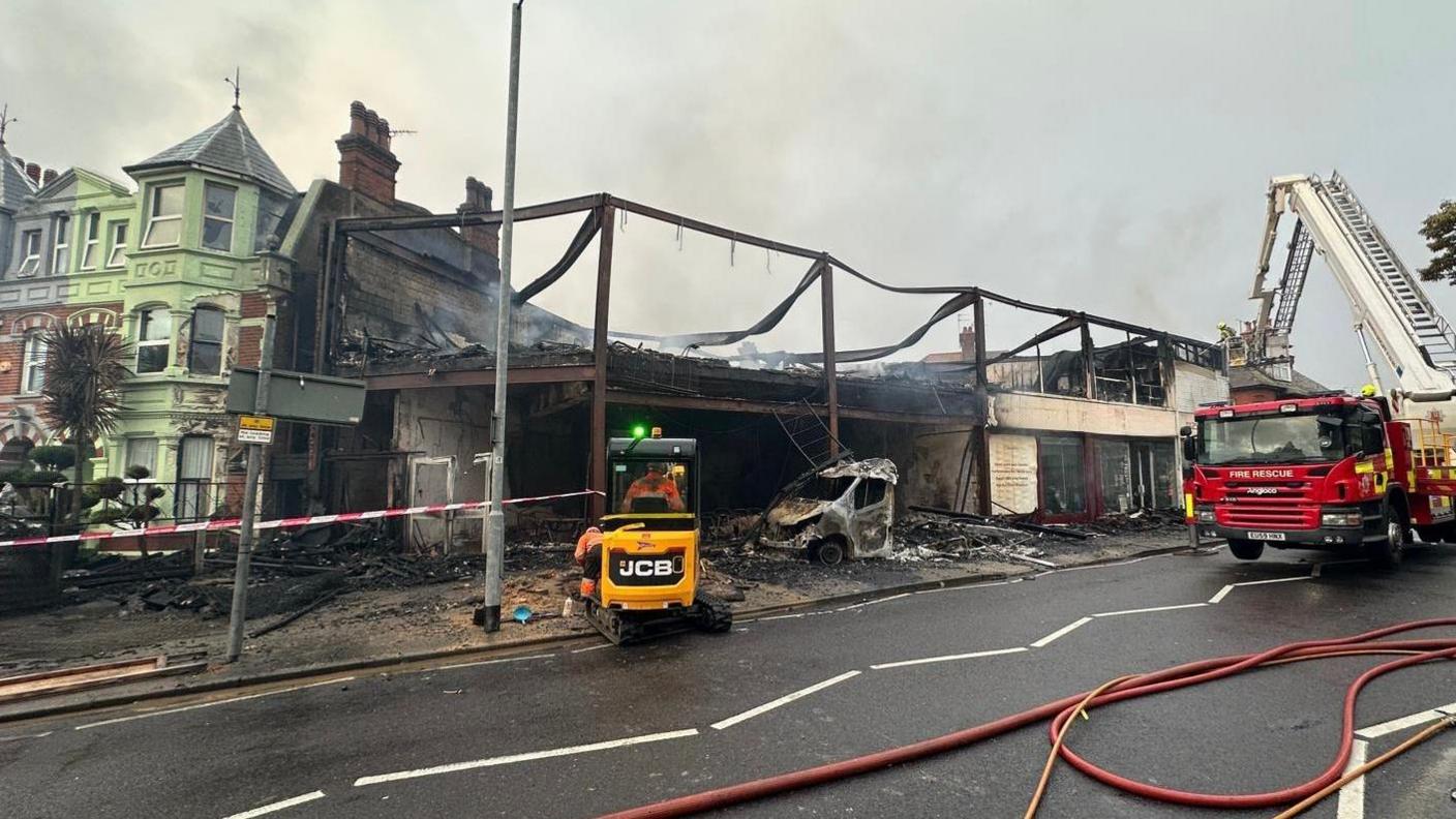 Smoking remains of the two storey buildings missing their walls and roofs. There is charred rubble on the ground and what looks to be the front of a car, which has also been burnt. A cordon is in place and a fire engine and yellow JCB are on the scene.