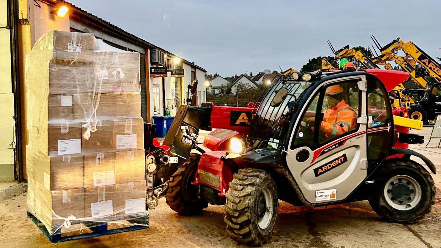 A forklift vehicle lifts a pallet filled with boxes that are wrapped in plastic packaging. A man wearing an orange high-vis jacket with an orange hard hat operates the vehicle. A warehouse can be seen behind the vehicle.