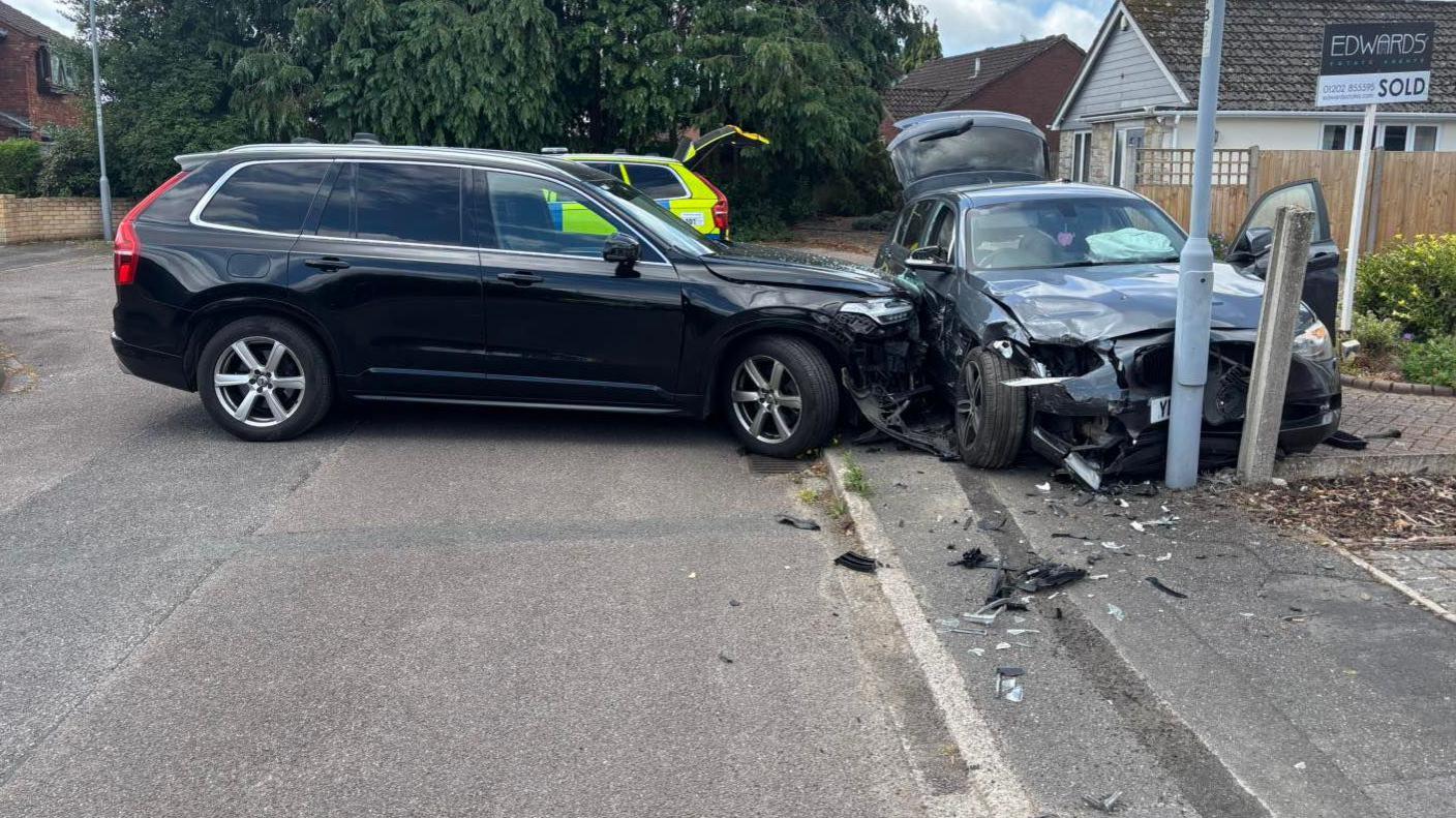 A car has rammed another car, leaving both badly damaged. The BMW has been shunted onto a pavement and has possibly hit a lamppost - its front and side are wrecked. The front of the unmarked police car is damaged. Another police car with markings is in the background.
