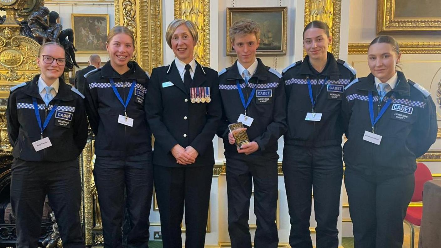 Six people wearing cadet and police unfiroms are standing in a line. They are smiling for the photo. A boy in the centre is holding a glass trophy, while a woman next to him has medals on her uniform. There are two girls either side of them.