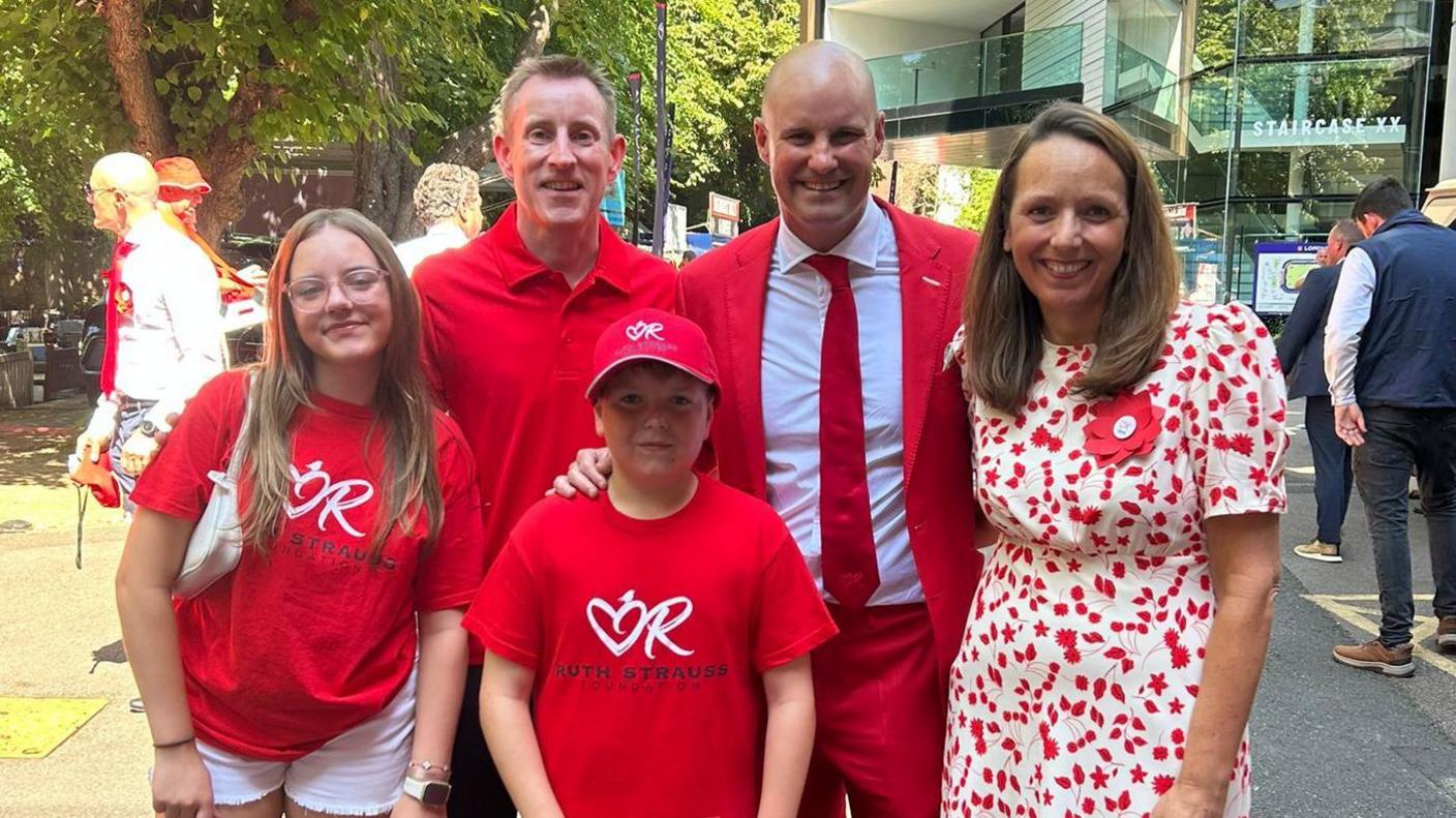 The family of four all dressed in red huddling around ex-England cricket player Andrew Strauss. Behind them is a glass building and a small patch of grass.