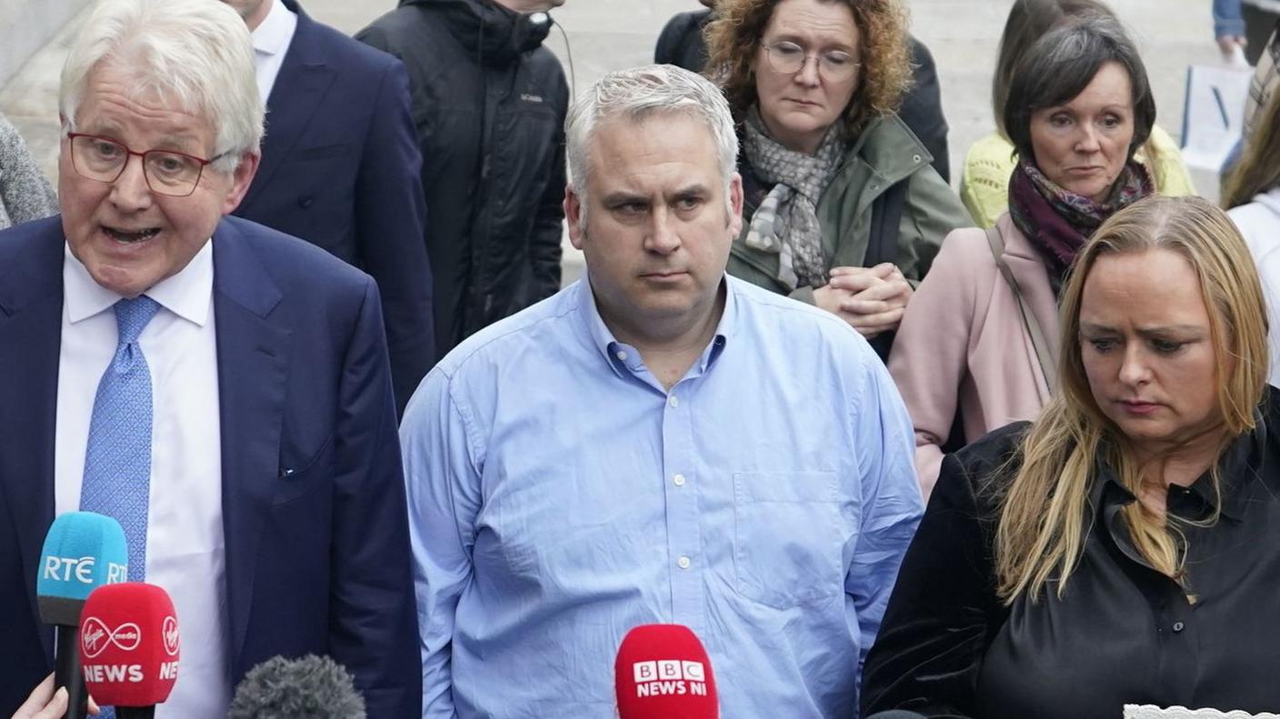 Hugh Harper, whose daughter, Leona, was killed in the blast is in the middle of the picture. He is wearing a blue shirt and has fair hair. He is looking into the distance. He is with picured with other bereaved families. ONe of them, a woman at the front, is holding up a picture of her loved one. Solicitor Damian Tanswey (far left) is wearing a dark suit, blue tie, white shirt and has glasses. There are microphones in front of the group as they speak to the media.