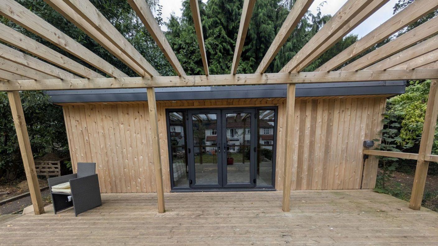 A wooden shed with a deck that has two glass doors and window next to each side of the door. A reflection shows a house on the opposite side and parts of the garden. There is a single grey chair in the corner of the shed.