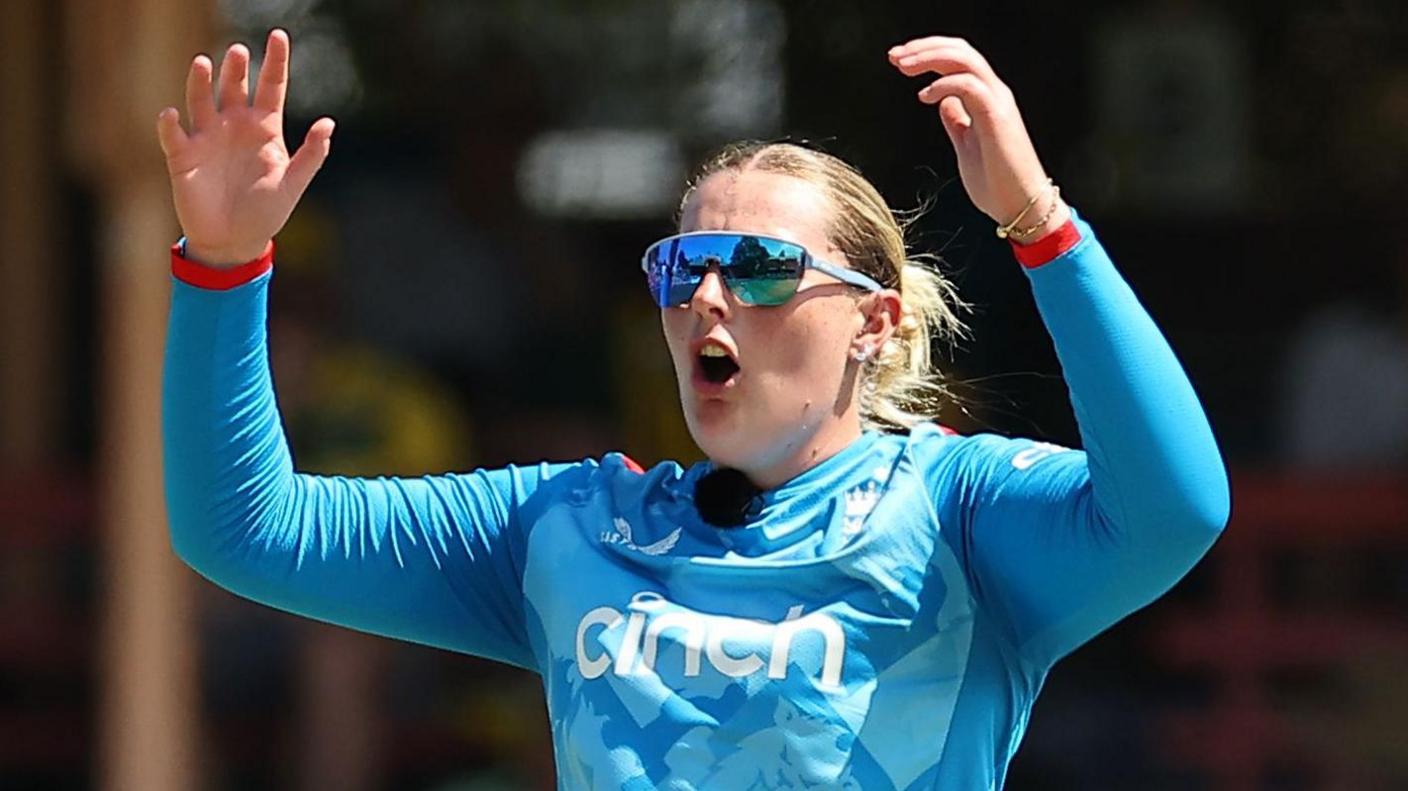 Sophie Ecclestone with raised hands, wearing an England limited-overs shirt and sunglasses