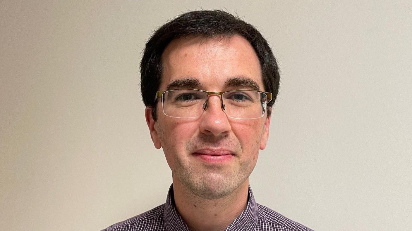 Dr Gavin McCluskey smiling at the camera as he stands in front of a beige wall. He has short black hair and is wearing thin framed glasses. He is also wearing a purple and white checked shirt.