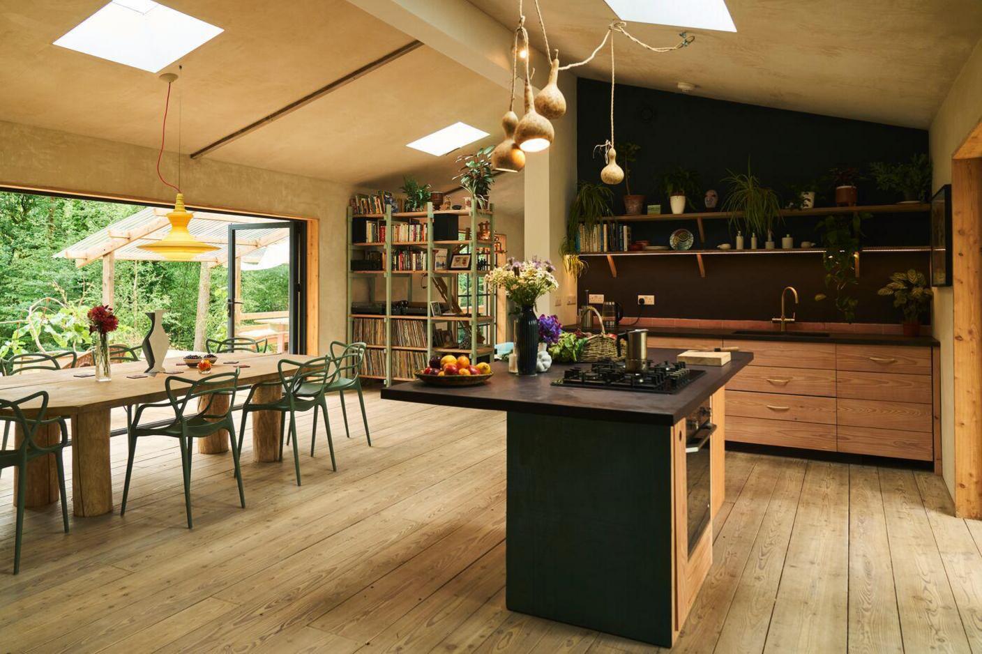 A shot of the interior of the house. A kitchen area which has wooden drawers and black counter top. In the kitchen there is shelving with plant pots, books and kitchenware. A bowl of fruit, flowers and cooker top sit on the counter of the island. Beside there kitchen there is a wooden table with green chairs and a book case.