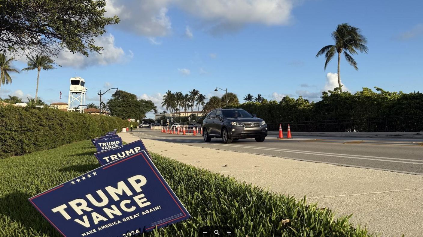 A car drives past Mar-a-Lago - there are Trump Vance campaign signs in the grass verge and a US Secret Service control tower is seen in the distance