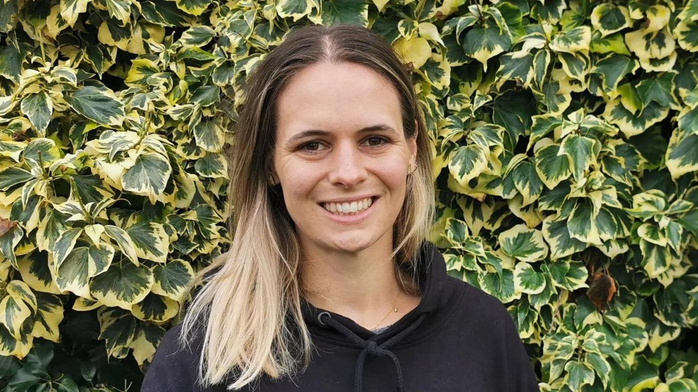 Helen Darragh is standing in front of a leafy bush, the leaves are green and yellow. She has long brown and blonde hair, is smiling at the camera and is wearing a black hoody. 