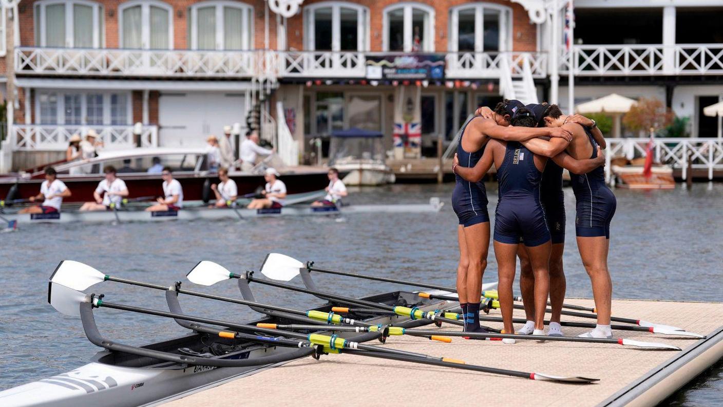 A team of four rowers in lycra huddling with their arms around each other's shoulders on a pontoon. The river and a row of boathouses can be seen in the background, and a rowing boat is moored next to the pontoon.