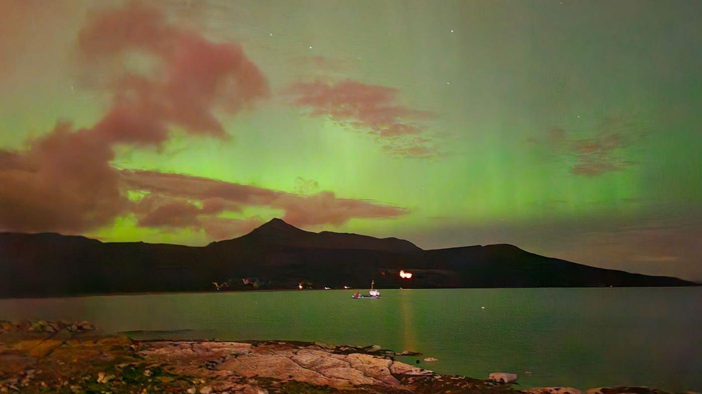 Green aurora behind hills and reflected in the sea.