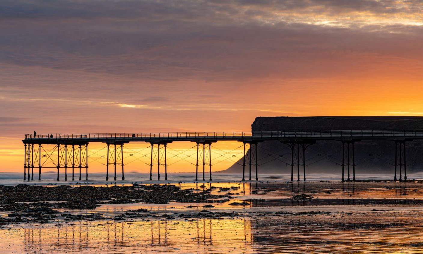 A pier juts out into the sea with a cliff and orange sunrise beyond, all reflected in the wet sand in the foreground.