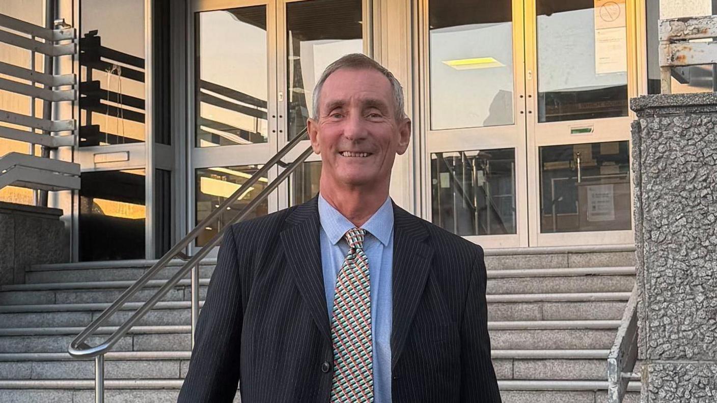 A man in a suit and tie smiling at the bottom of wide grey stairs. Two sets of glass doors to a grey building are at the top of the stairs.