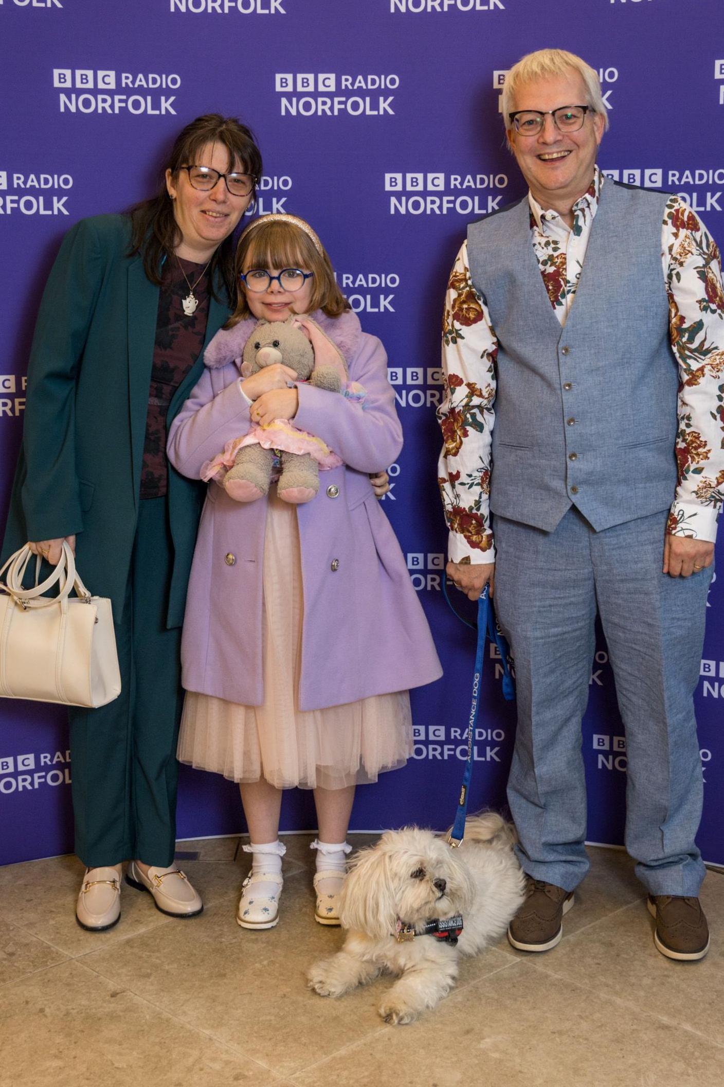 A man, a woman, a young girl and a white fluffy dog pose in front of a purple BBC Radio Norfolk background. The girl is holding a stuffed toy and is wearing a pink dress and purple coat. 