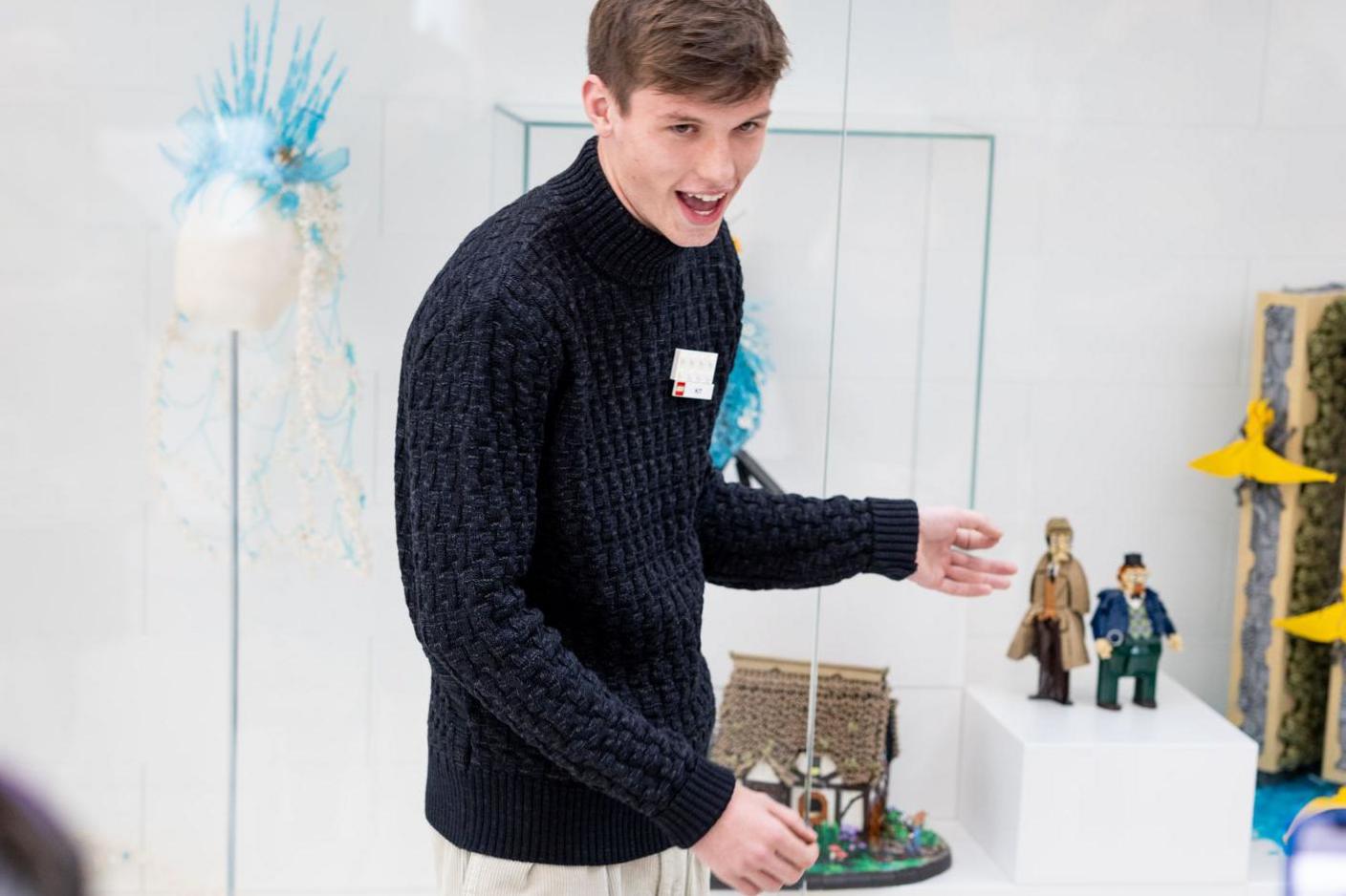 Kit Nugent, who has brown hair and is wearing a navy jumper and beige tousers, points to his models in a glass case during the opening of the exhibition at the Lego House.