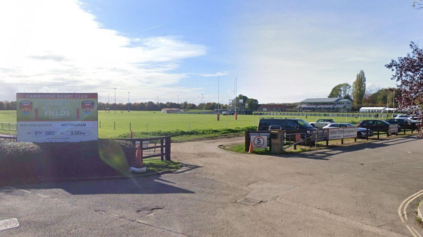 A view of the entrance to the Cambridge Rugby Football Club. A large sports field can be seen just beyond a small car park, in which cars are parked. 