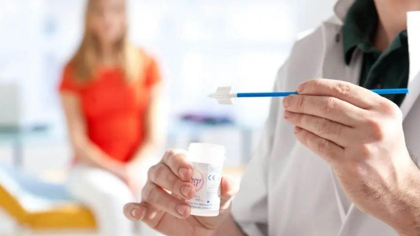 A doctor in white scrubs holding a cervical smear test. A woman in white trousers and an orange top sits behind her, blurred out in the background.