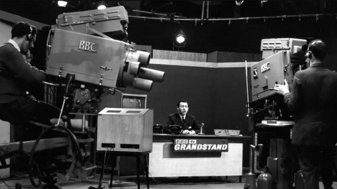 A black and white archived image of David Coleman sitting behind a desk in a filming studio set. The desk says BBC TV Grandstand on the front, and above him are studio lights suspended from metal bars. In the foreground there are two men operating massive cameras on stands.