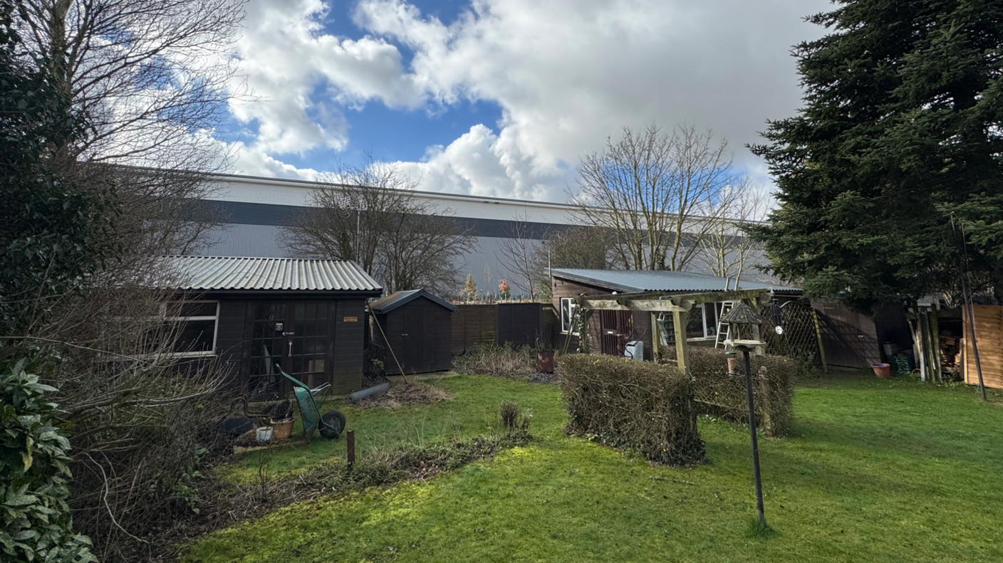 A large warehouse can be seem backing onto a back garden in which there are sheds, bird feeders, a wheelbarrow, and other gardening equipment.