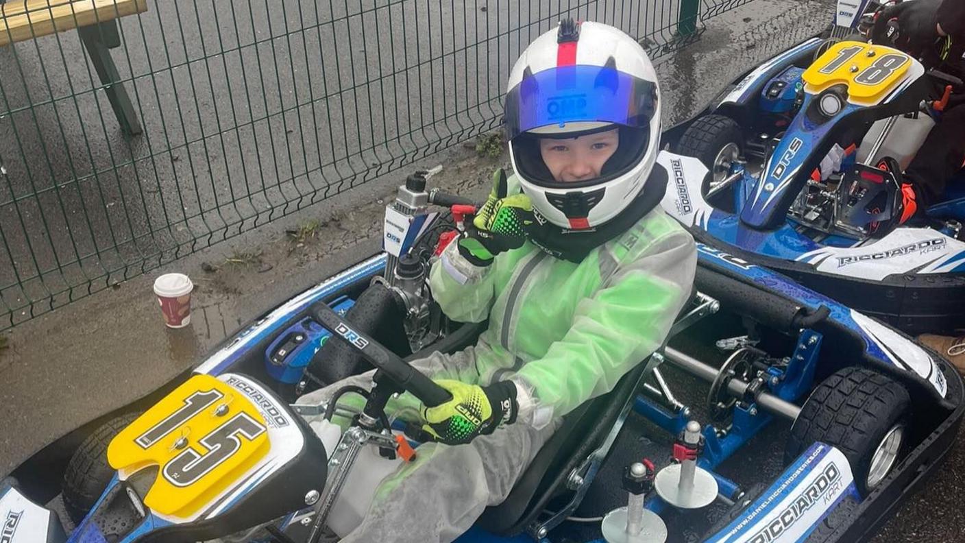 Jacob Kent in a green racing suit sits in a blue go-kart with his thumbs up