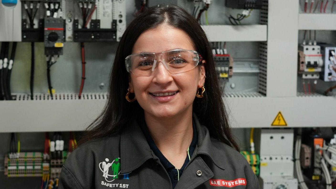 A woman with dark hair and wearing safety goggles and a black shirt with the BAE Systems branding on it. She's smiling at the camera and there's a wall of electrical equipment in soft focus behind her.