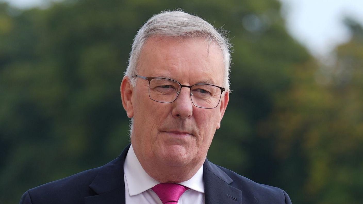 A man with white hair and dark rimmed glasses looks off to the side of the camera. You can see the top of his shoulders, he's wearing a navy blue suit, a white shirt and a bright pink tie. The background is blurred, but you can make out trees and greenery.