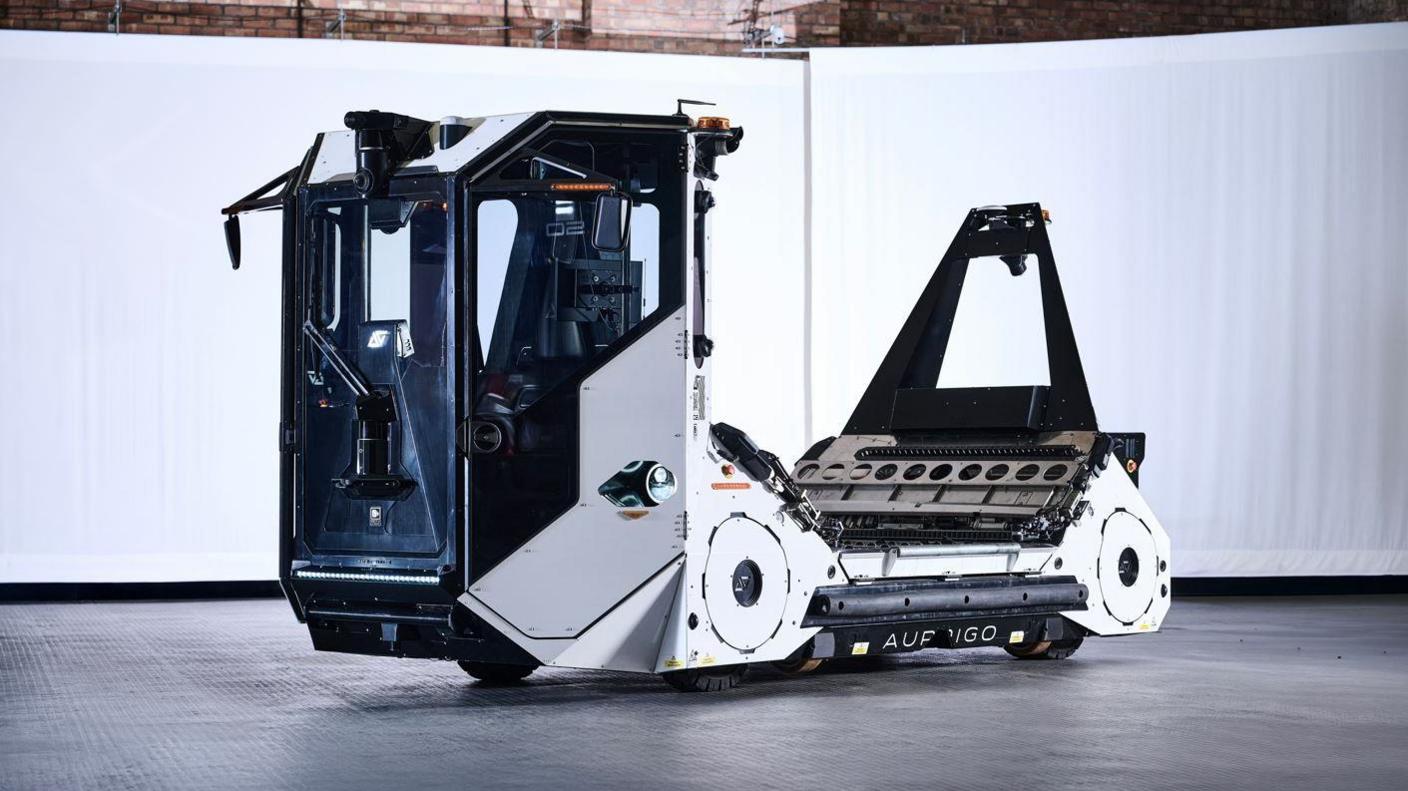 The Auto-DollyTug. The vehicle is white with a large, windowed cabin at the front. The rear of the vehicle is a platform where luggage can be stored.