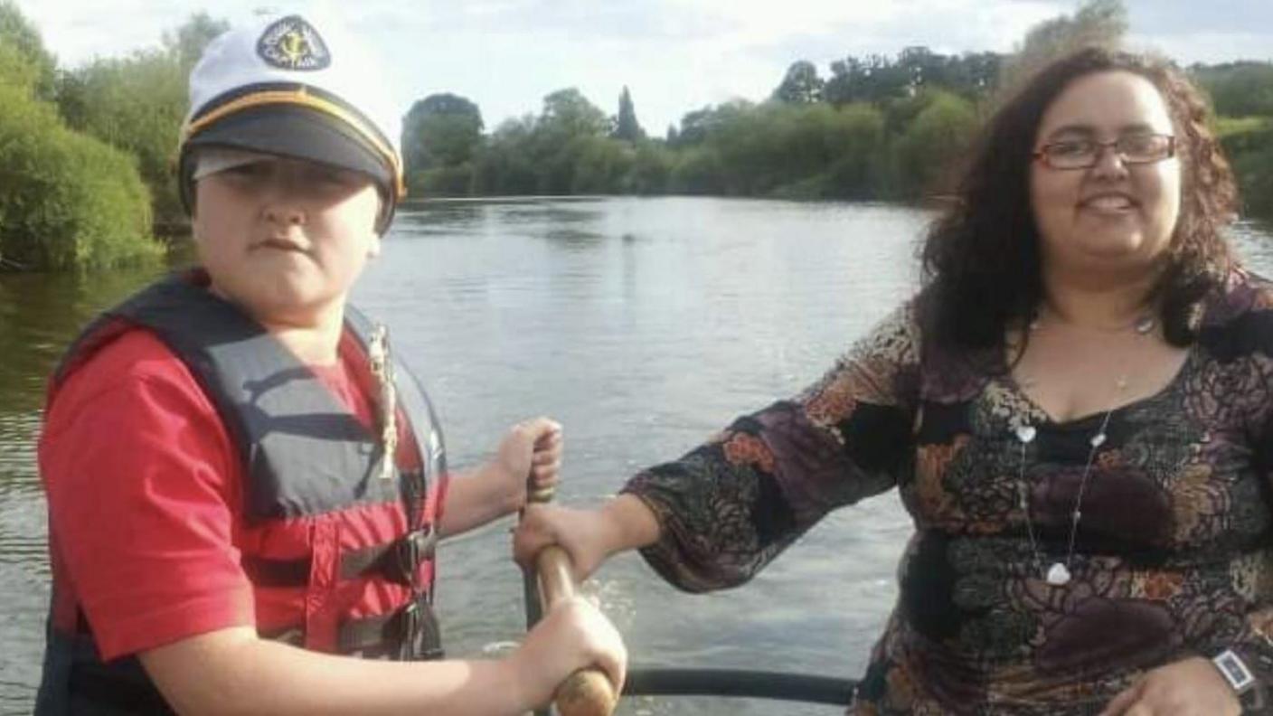 Tomas Cozens as a young boy with his mum Jo. They are on a boat on a river and are holding a wooden handle that steers the boat. Tomas is wearing a red t-shirt and a black and red life jacket with a white, Navy-style captain's hat. His mum has curly dark hair and rectangle-shaped glasses with a dark, patterned long-sleeved top on. The river and trees are visible in the background