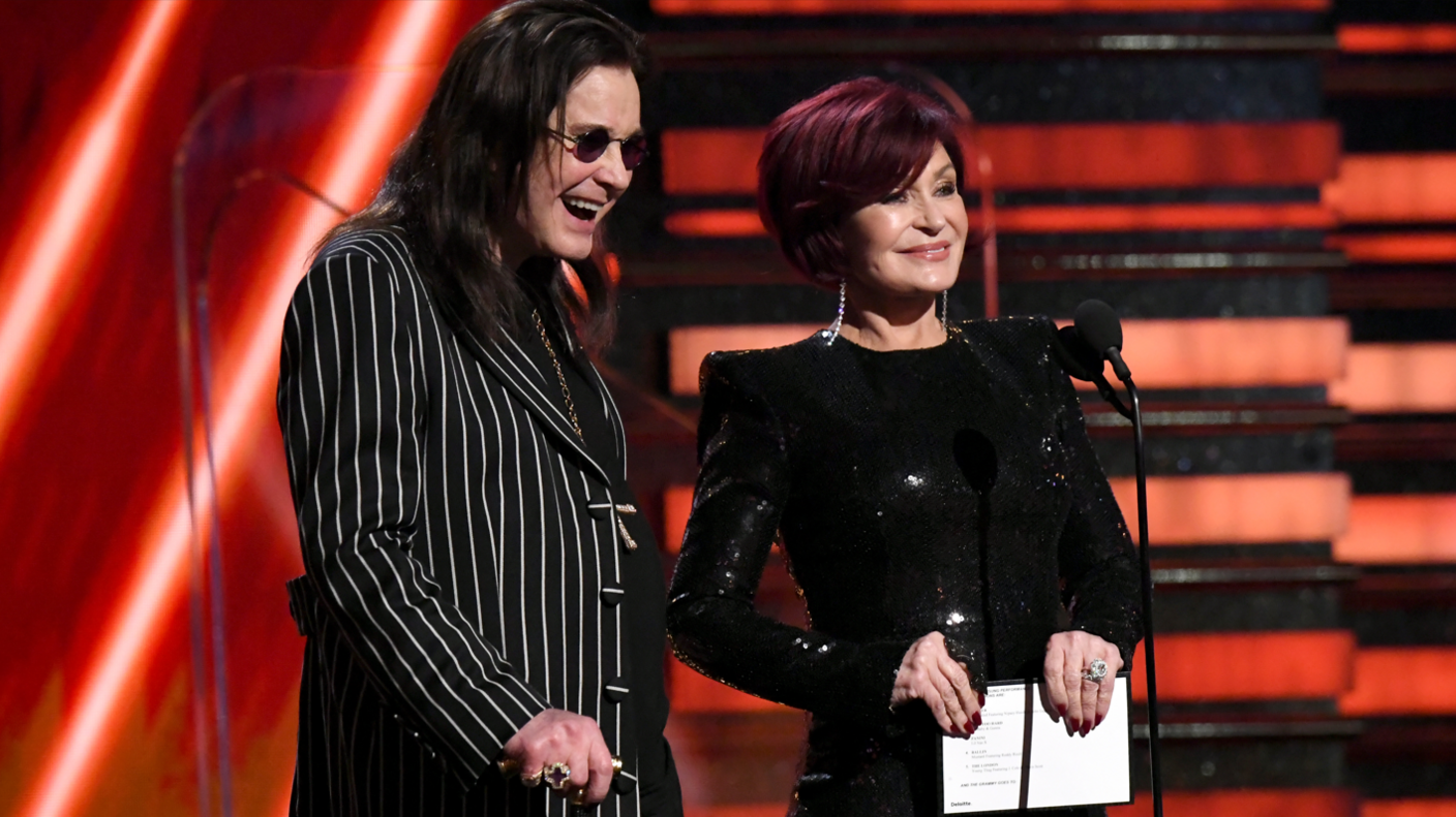 Ozzy and Sharon Osbourne smiling on stage at the 2020 Grammy Awards: Ozzy stands on the left wearing a black and white striped jacket, a cross-shaped ring, and black sunglasses. Sharon wears a shiny black dress and holds a piece of paper, with diamond rings visible on her ring finger