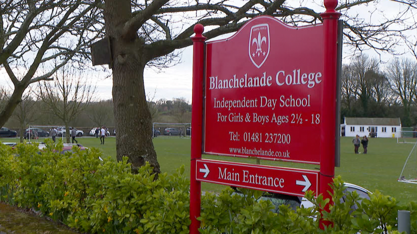 A wide shot of the college's signage. It reads "Blanchelande College, Independent Day School for girls and boys". The sign is red with white writing. A school playing field can be seen behind.