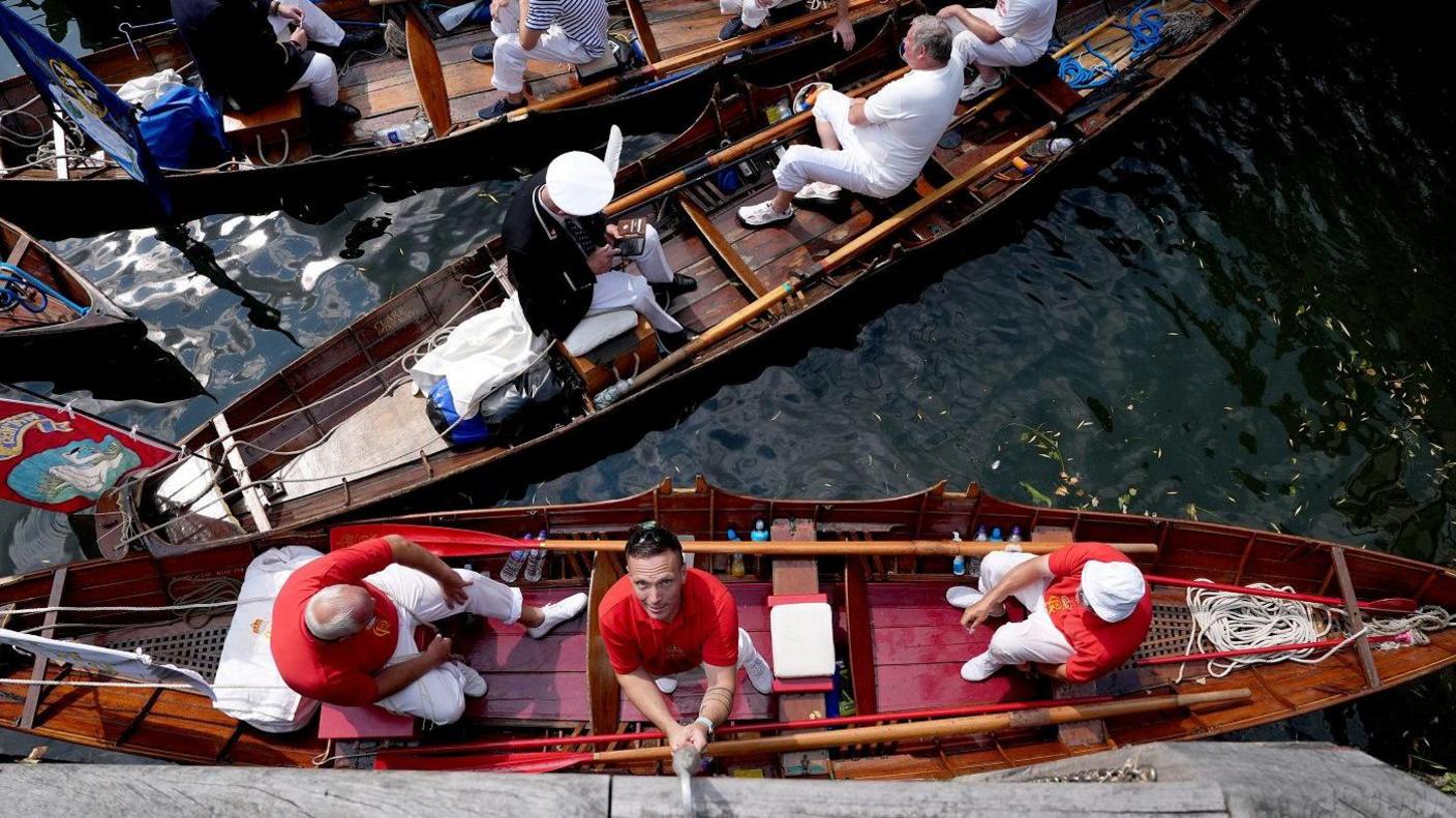 An overhead view of several wooden rowing boats floating on a calm body of water, with groups of people dressed in red and white nautical attire. The boats are moored closely together, and one man is reaching up towards the bank.