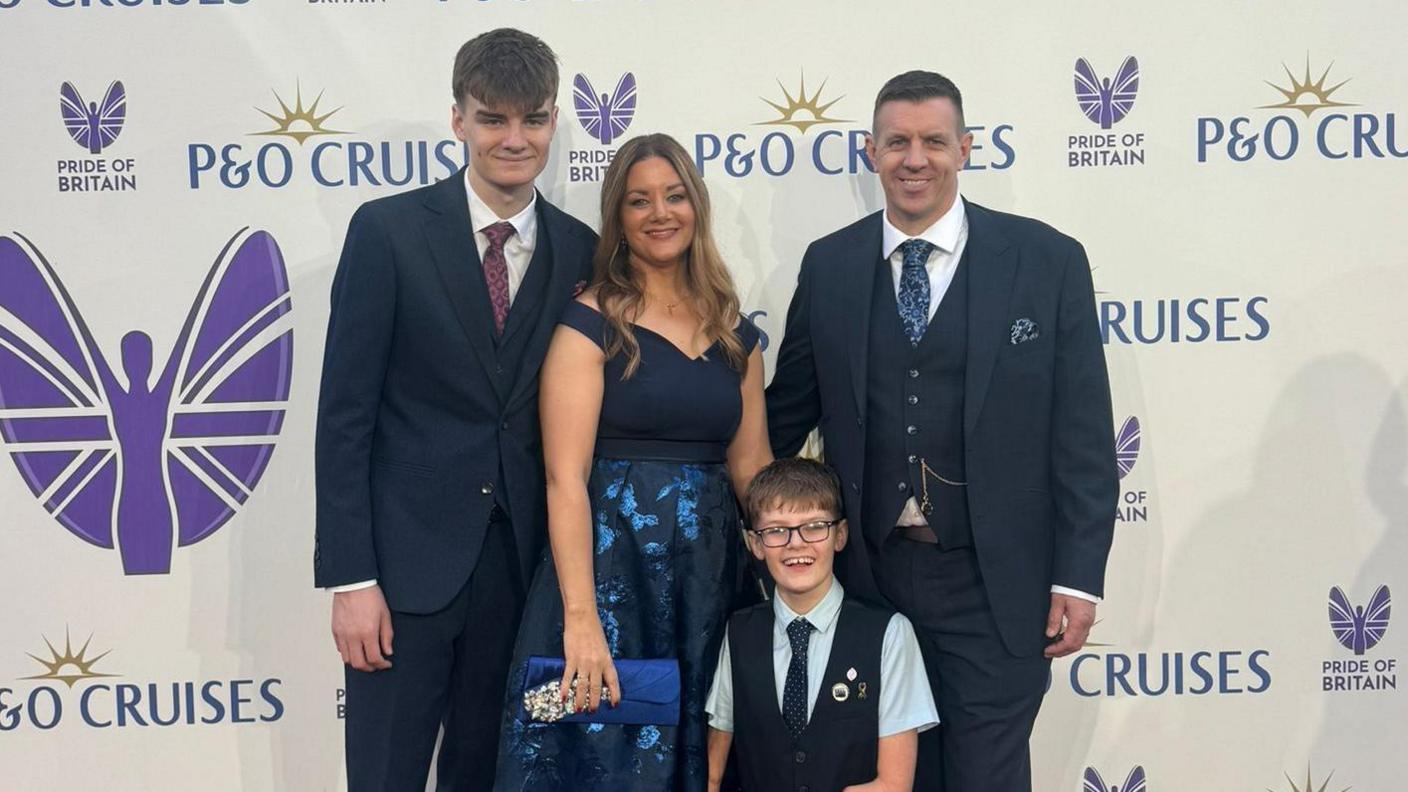 A group photograph of the Mortimer family. Luke's older brother stands on the left of the group, he is wearing a dark suit, white short and tie. Luke's mother is standing in the centre of the group. She has long, light brown hair and is wearing a blue dress. On the right of the group is Adam Mortimer. He is wearing a navy, three-piece suit with white short and blue tie. Luke is at the front of the group. He is wearing a white shirt and a navy wasitcoat.