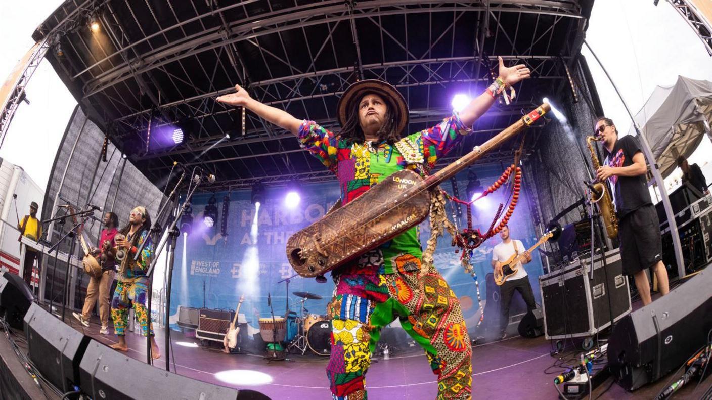 A performer on stage wearing a colourful shirt and trousers in a traditional African print. He has long dreadlocks and is facing the crowd with his arms outstretched wide. Hanging off him is a kora, a traditional African instrument, which looks like an elongated wooden guitar.