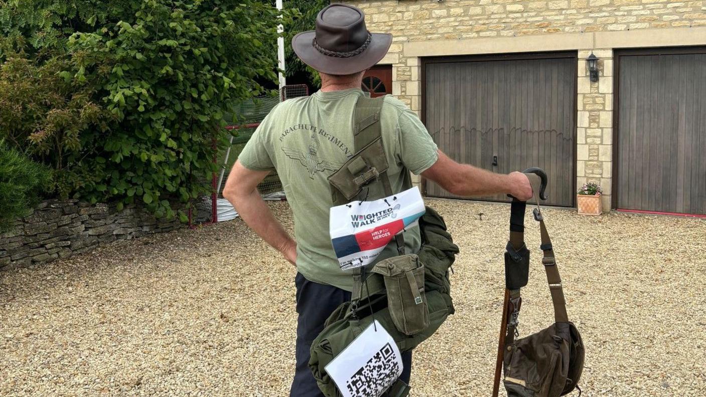 Philip with his back the camera on a driveway wearing his old army gear and a leather brimmed hat with a help for heroes sign on his back