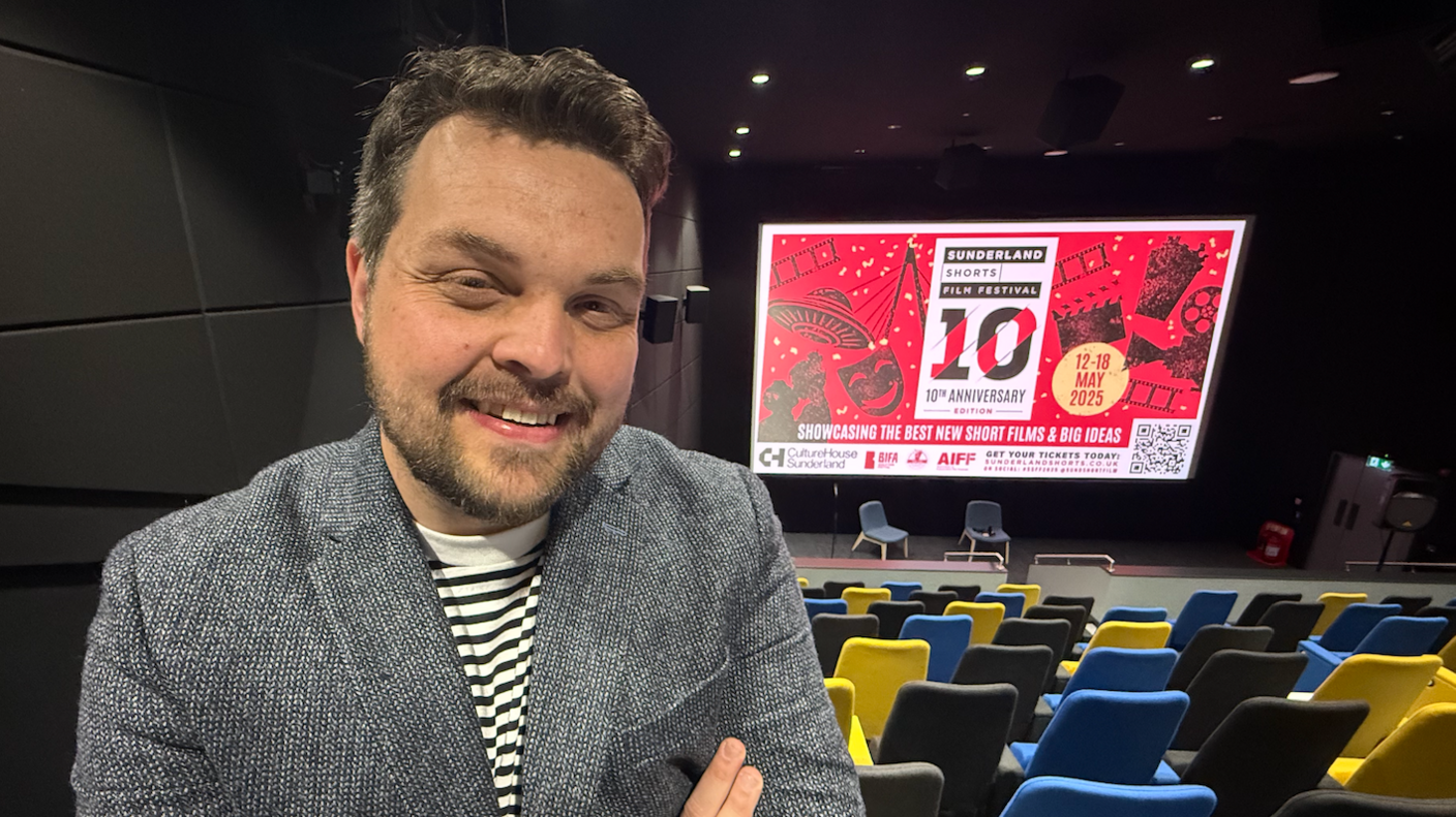 Chris Allan wearing a black and white striped shirt and a grey patterned blazer stands at the top of the auditorium, which has black, blue and yellow seating, while the cinema screen displays a red and white-coloured graphic showing the film festival is in its tenth year.