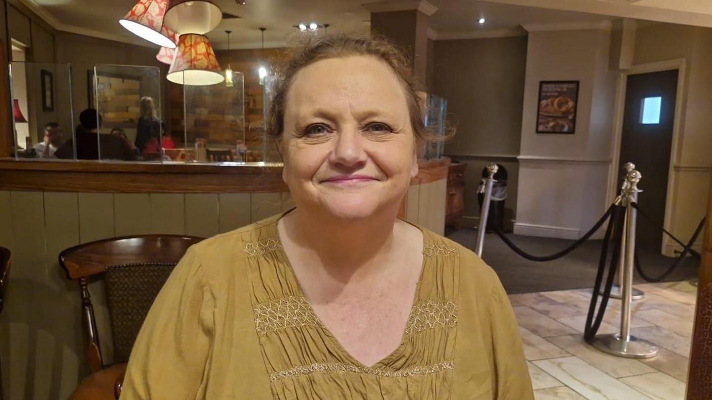 Wendy Goldie in a gold top looking at the camera in seating area