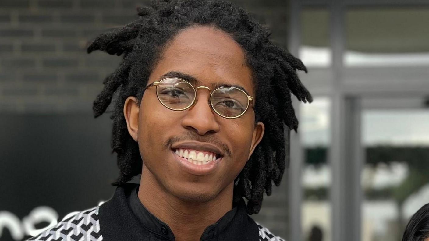 A man outside a grey brick building, looking and smiling at the camera. He has dark skin, black dreadlocks, and a patterned white and black polo shirt. He is shown from the shoulders up.