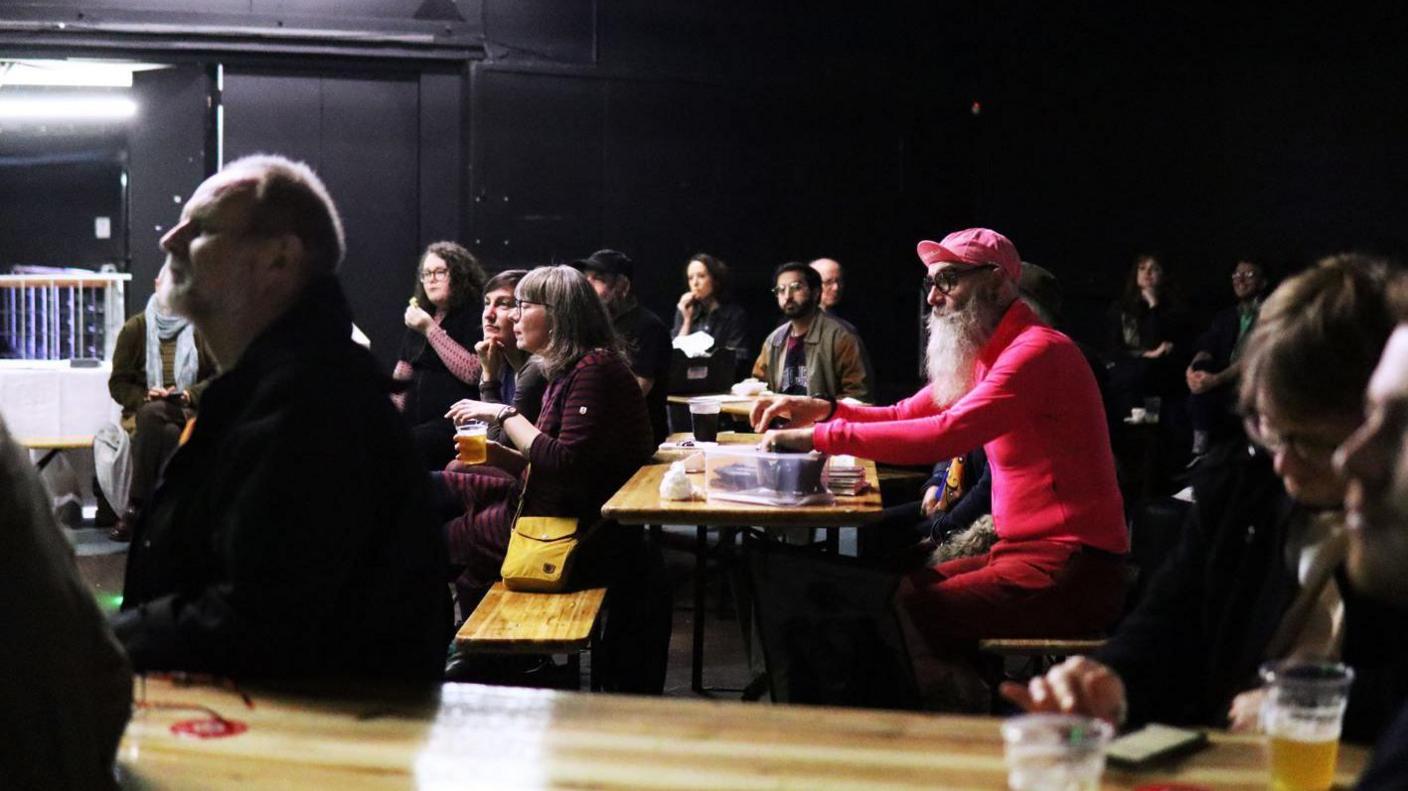 A group of dozens of men and woman sat down on benches in a dark room.