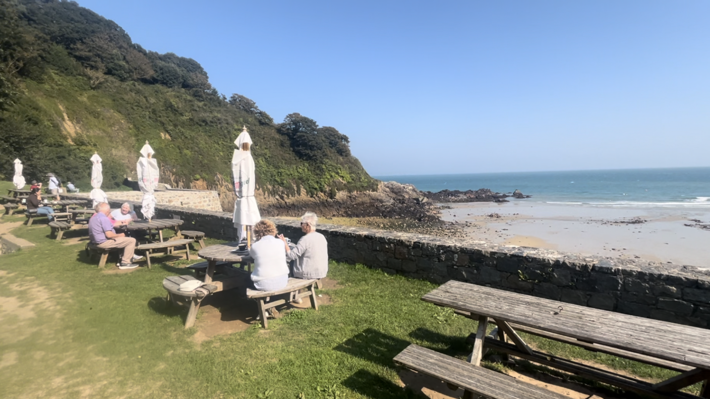 Several wooden benches and tables on a grass patch overseeing a pebbled beach on a sunny day. A handful of people sit at the tables.