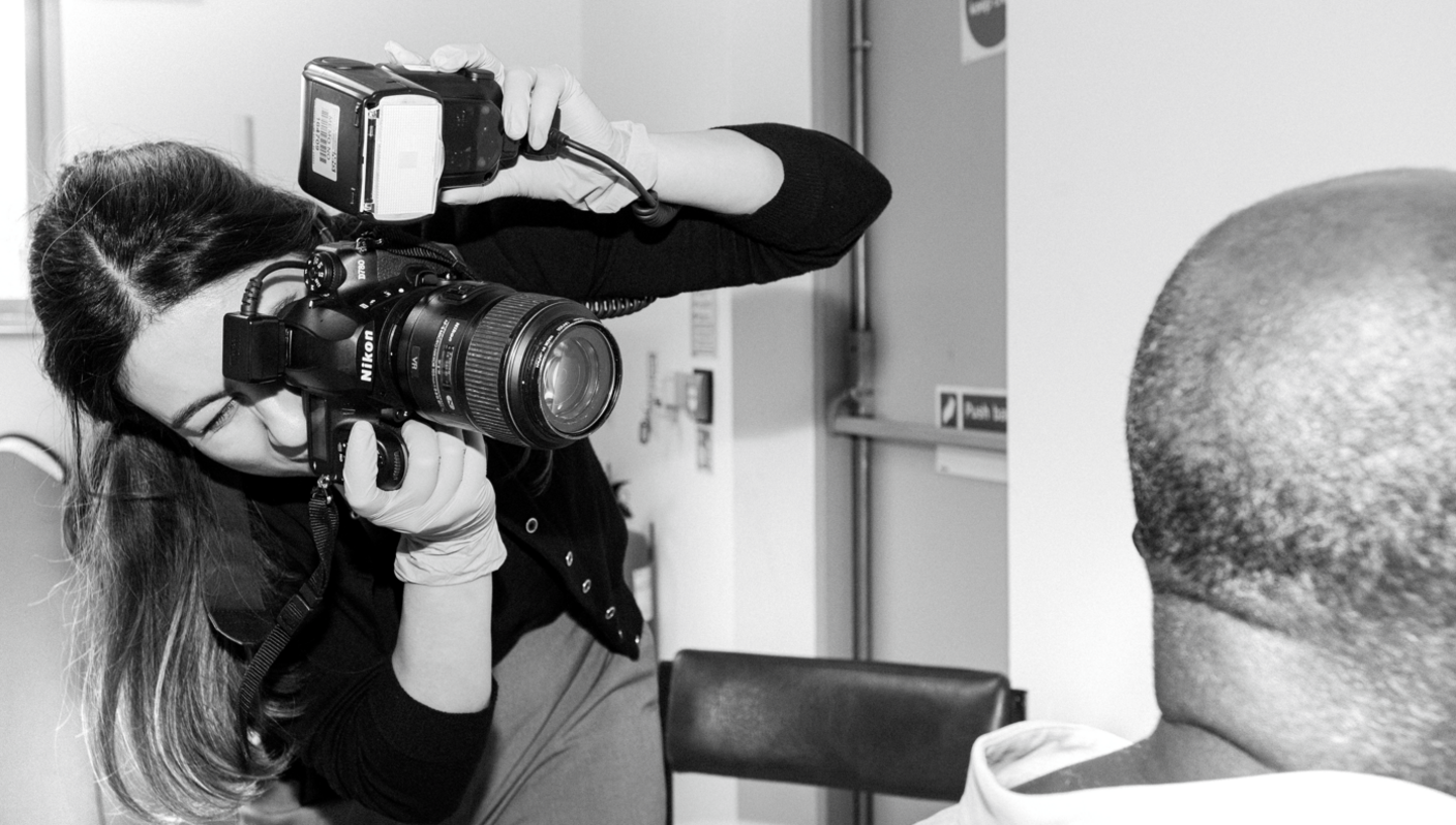 A black and white shot of a female photographer holding a large camera and taking a picture of a seated man, who has his back to the picture