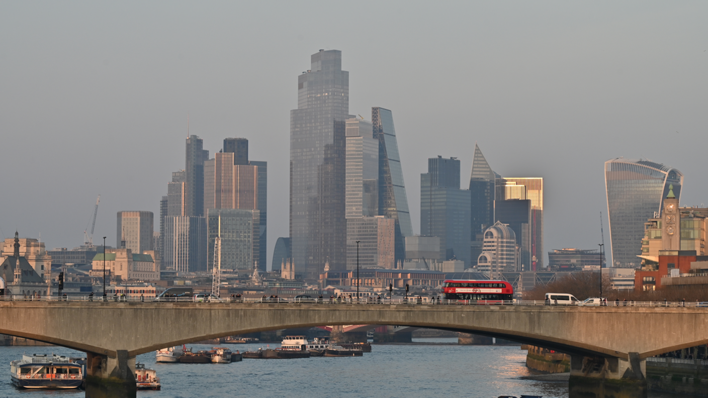 Vehicles travelling across Waterloo Bridge with the towers of the City of London seen behind it and the vessels on the River Thames beneath it