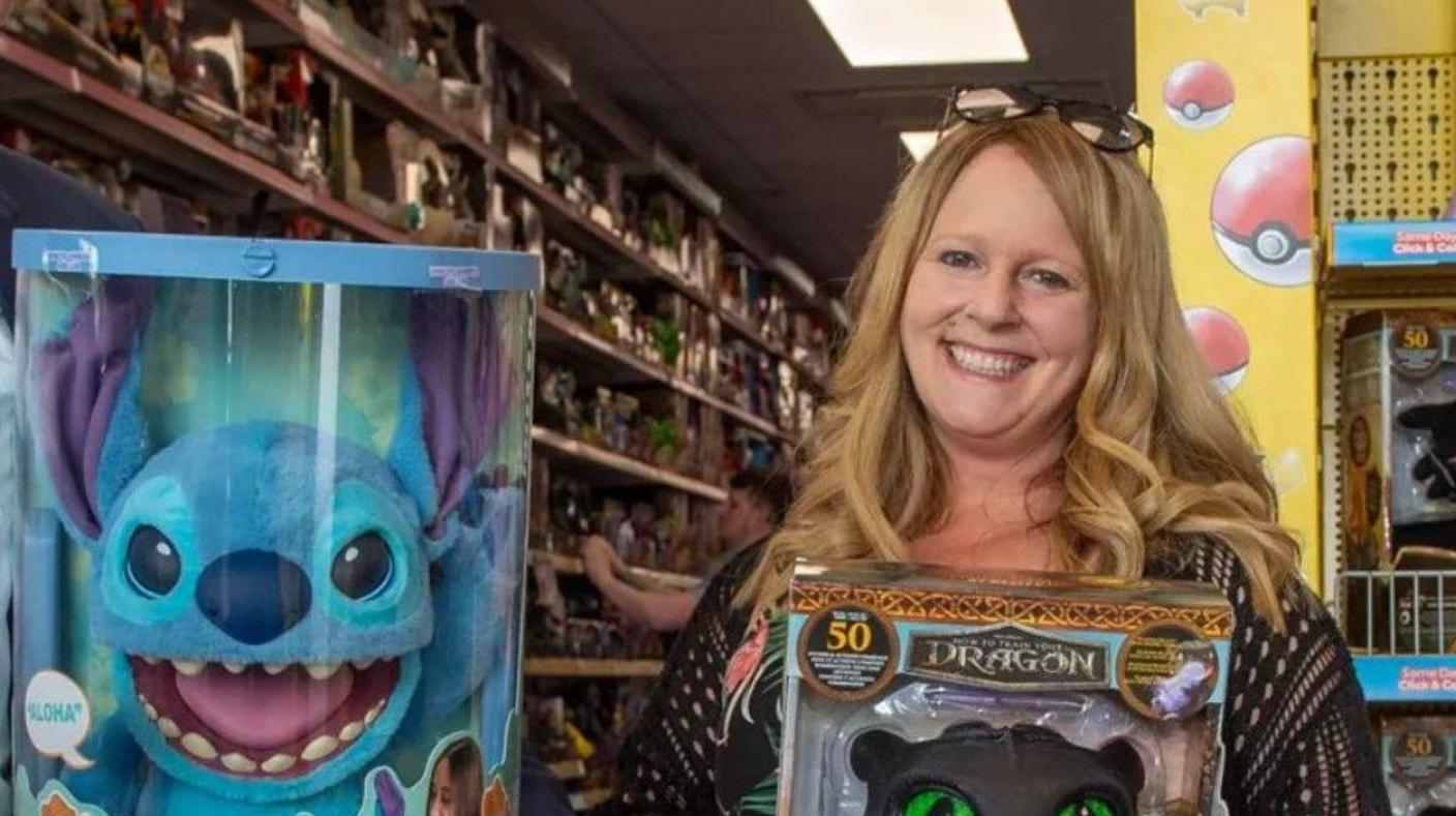 A woman holding a toy and smiling. Another toy is prominent on the photo to her left. Toys are on shelves in the background.