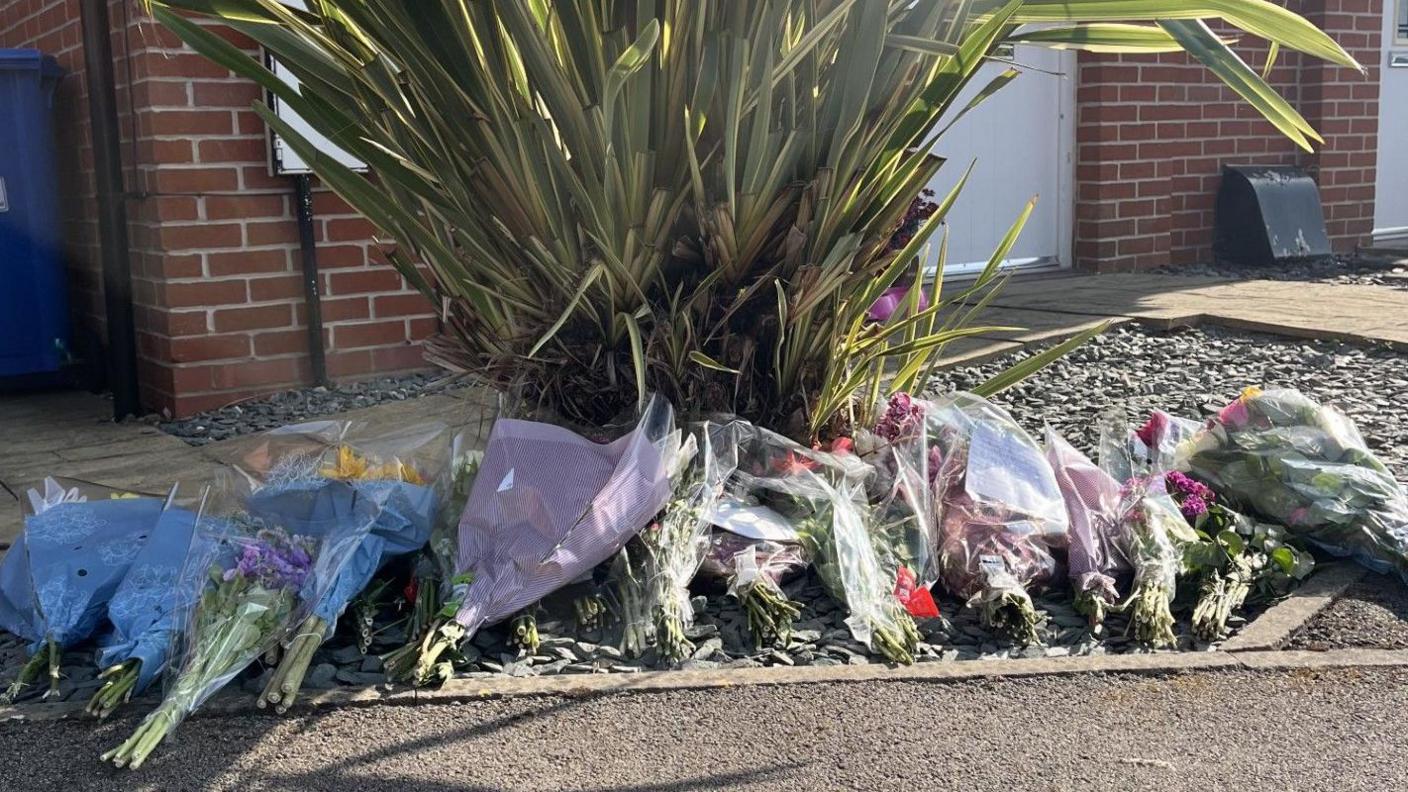 A row of floral bouquets , some in blue wrapping, are laid out in front of a large ornamental grass. In the background is the wall of a brick-built house with a white door.